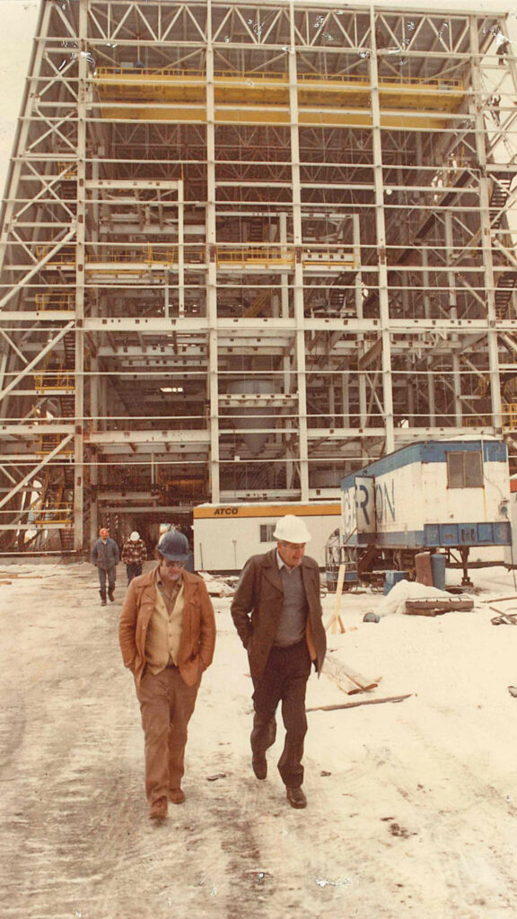 4 people walking through a large construction site in 1970s